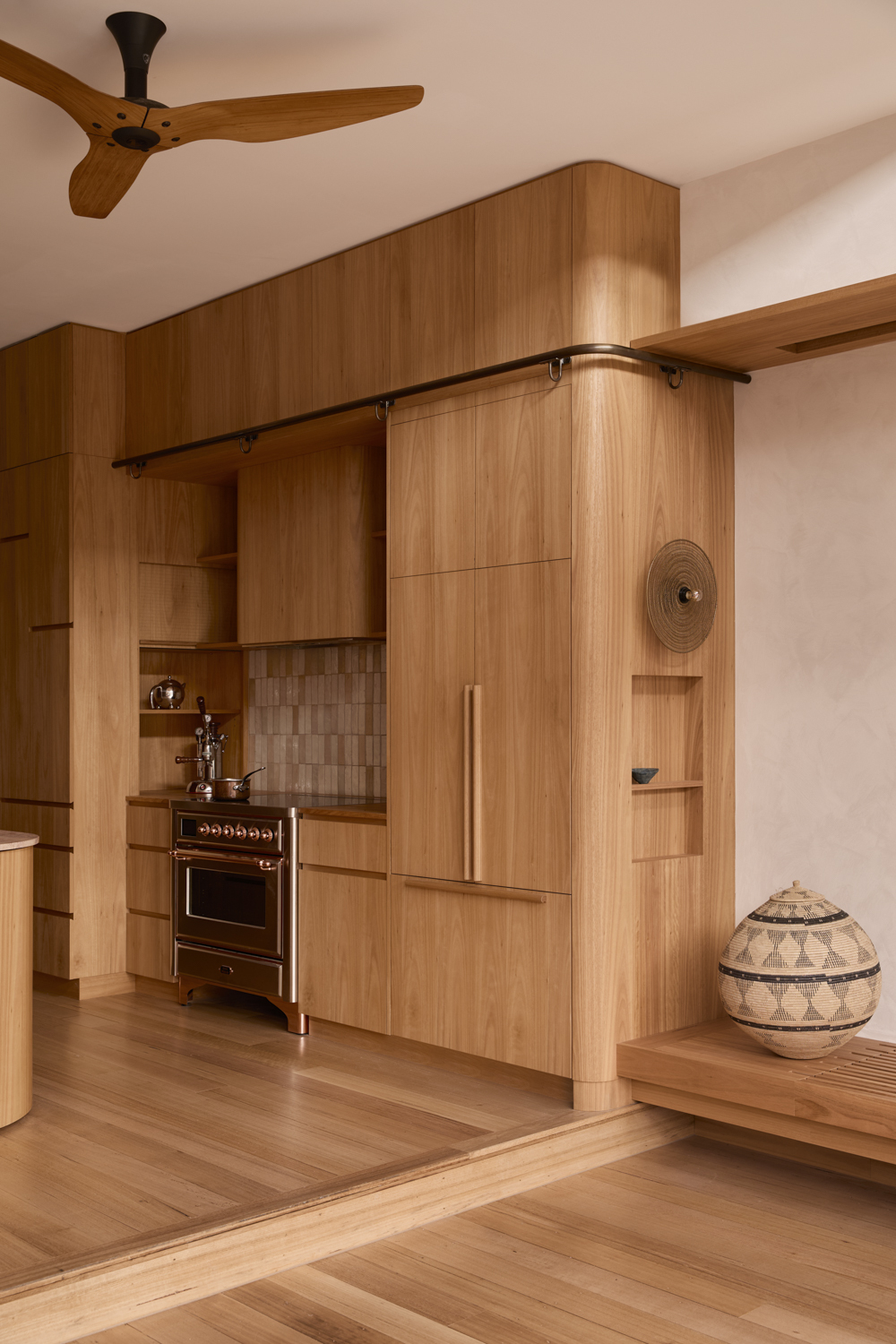 A wood-clad, open-space kitchen with a pale, geometric surface of curved and straight lines, a tiled backsplash in ceramic rectangles, and brass detailing, illuminated by sunshine.