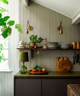 A traditional kitchen with open shelving, brass wall sconce lights and a green glass lamp on the counter