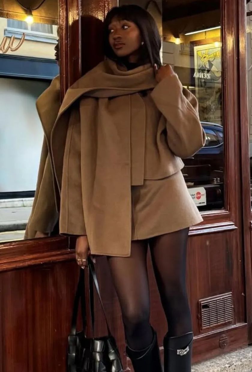 A photo of @emmanuelleuk's outfit, featuring a brown skirt co-ord with a scarf jacket and black boots, standing outside a patisserie