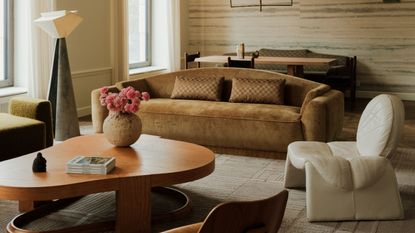 Image of a modern living room with a neutral rug, a velvet caramel sofa, a white leather accent chair, and an abstract wooden coffee table.