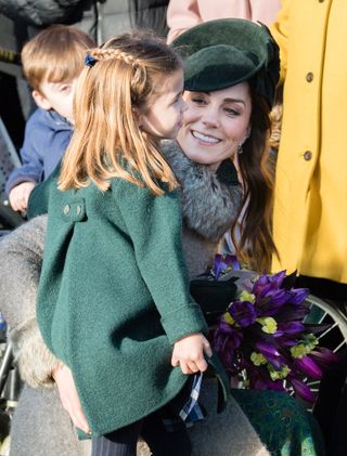 Princess Kate wearing a gray coat and green hat holding Princess Charlotte, who is wearing a green coat and braided hair