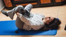 woman in grey tracksuit bottoms and relaxed top lying on a blue exercise mat in a figure-four stretch on tiled floor. 