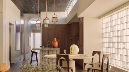 Image of an open kitchen and dining area with white walls, glass block windows, a round dining table, and wooden chairs. There is a marble island with three pendant lights hanging over it.