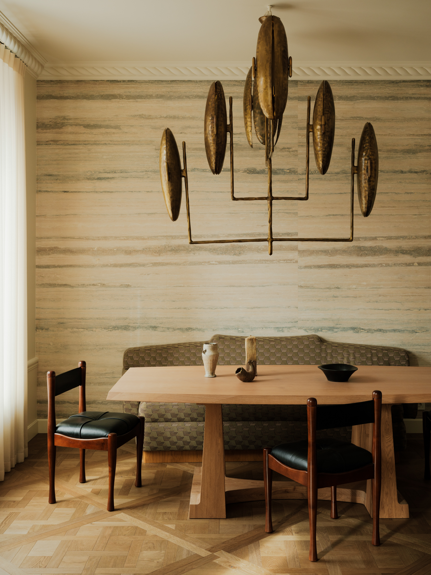 Image of a warm, contemporary dining room with a dark beige color scheme. There is a wooden dining table with a fabric bench seat against the wall and wooden and black leather chairs on the other side. There is a sculptural light fixture and textured wallpaper on the walls.