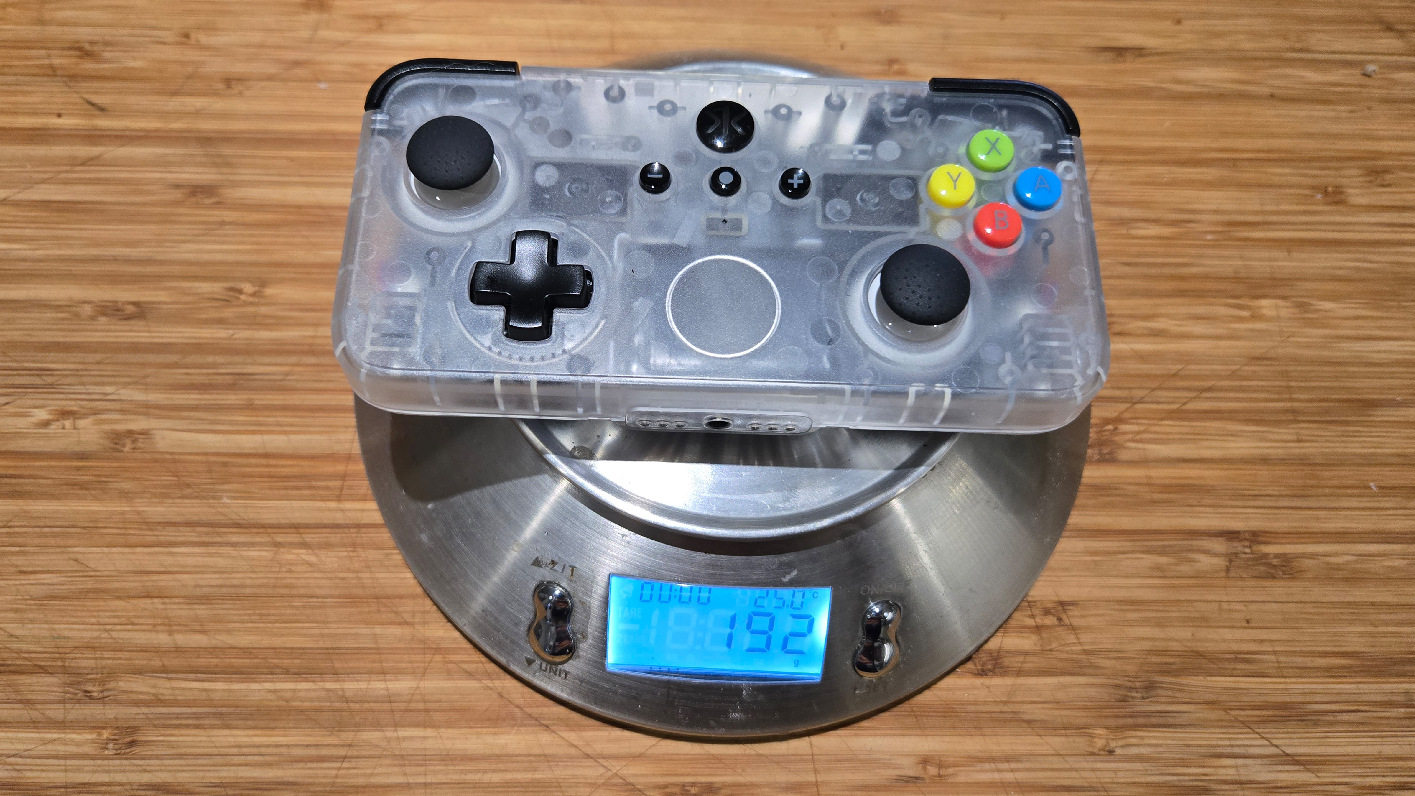 A Classic Clear edition of the CRKD NEO S rectangular controller sits on a small kitchen scale. The digital read out on the scale reveals the tiny controller weighs only 192 grams. The body of the controller is made of a cloudy clear plastic allowing the viewer to see the internals.