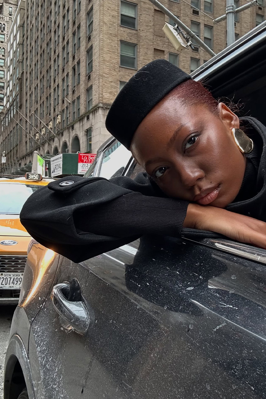 West Village winter trends are shown in a close selfie photo of a woman leaning out the window of a black car driving down a street in New York, wearing her winter outfit. She's wearing a black pillbox cap, oversized gold earrings, and a black coat.