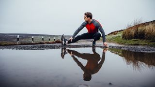 Male runner training outdoors in wet weather. His reflection seen in a puddle of water before him. He stretched his leg