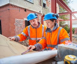 man and women in orange hi vis coats and blue hard hats on building site looking at plans