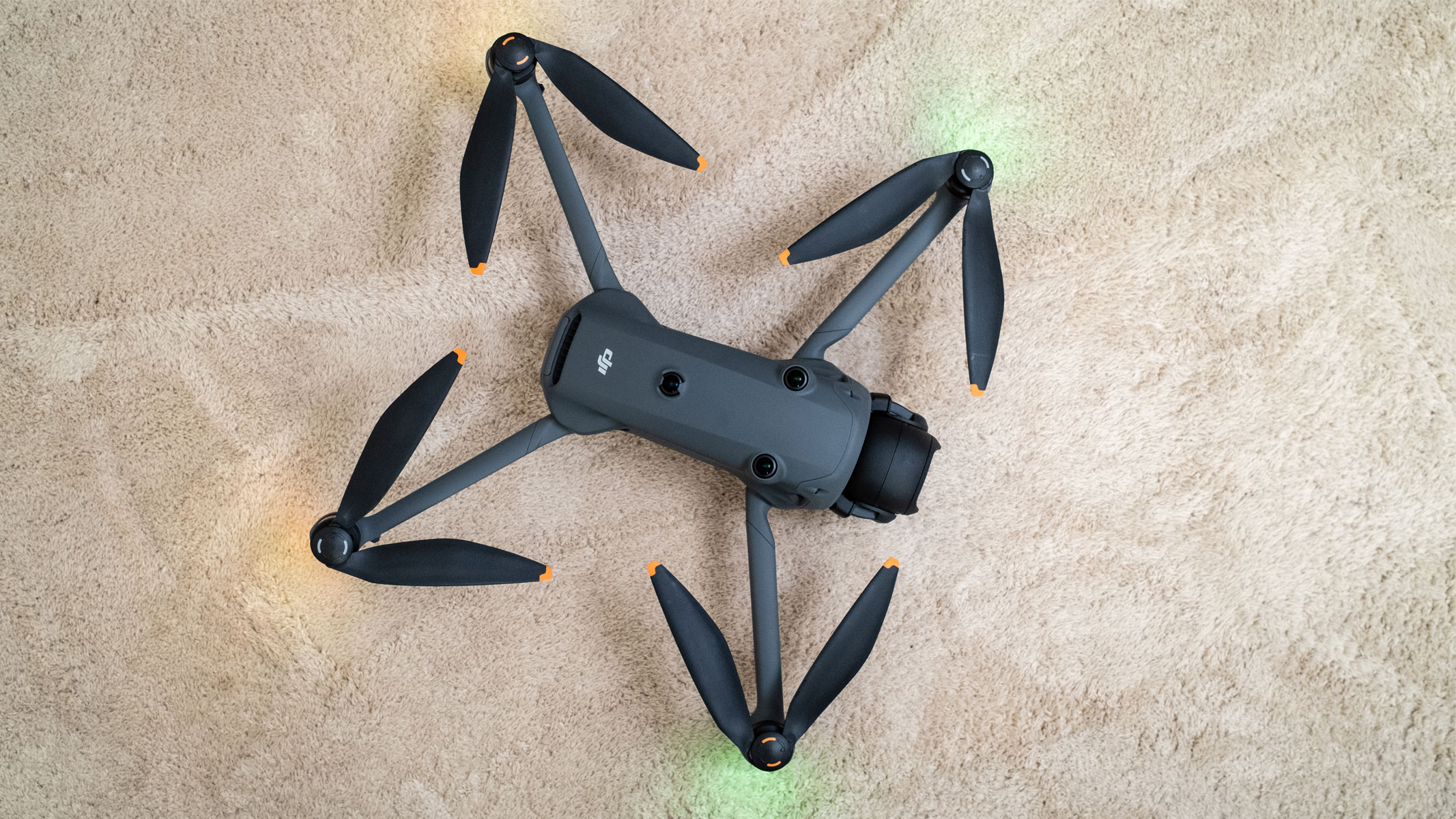 A grey drone has four propellers extending out of each corner of the central body with green lights shining from the front two propellers on a beige carpet.