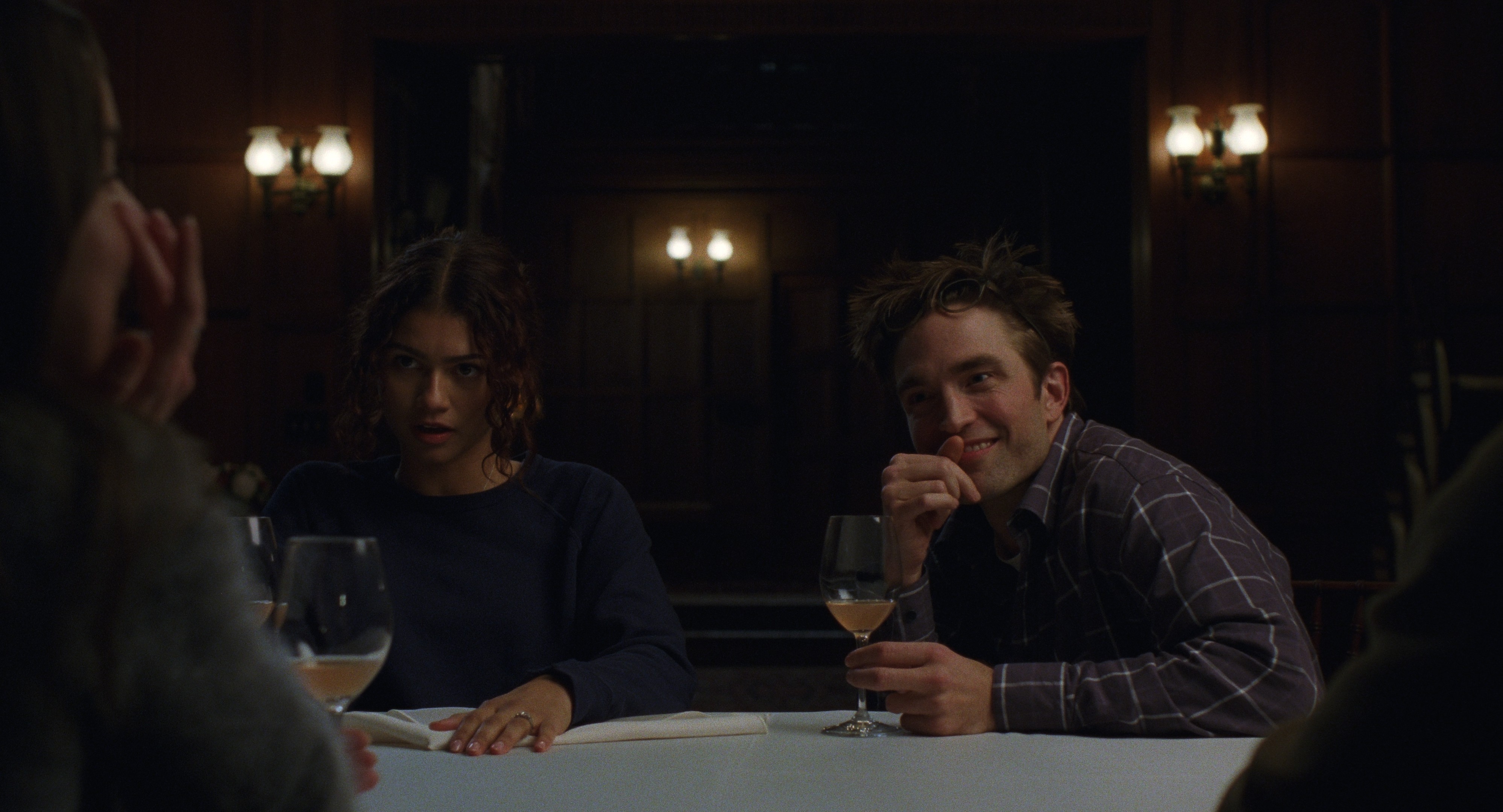 A young couple talking to friends around a dimly lit tables with a glass of wine at hand.