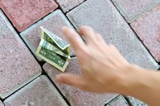 A hand reaches down to pick up a dollar bill left on the ground.