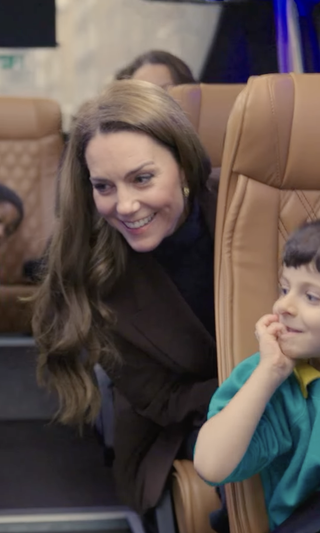 Kate Middleton wearing a brown blazer sitting on a bus seat and smiling leaning into the aisle with a little boy in front of her