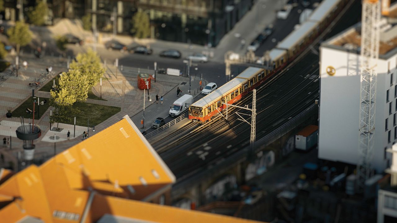 A red and yellow train navigates tracks near a busy urban plaza filled with trees and parked cars, framed by modern buildings, appearing as a miniature world as foreground and background are unsharp, showcasing a tilt-shift effect