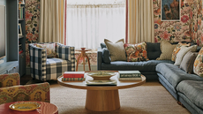 a heritage living room with bold, patterned wallpaper, a large blue sectional with many pillows on it, a gingham printed accent chair, a round coffee table, and a large window in the background covered by drapes