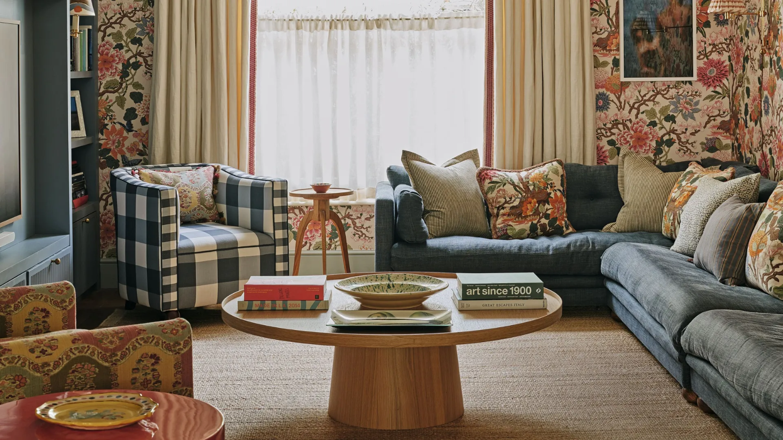 a heritage living room with bold, patterned wallpaper, a large blue sectional with many pillows on it, a gingham printed accent chair, a round coffee table, and a large window in the background covered by drapes