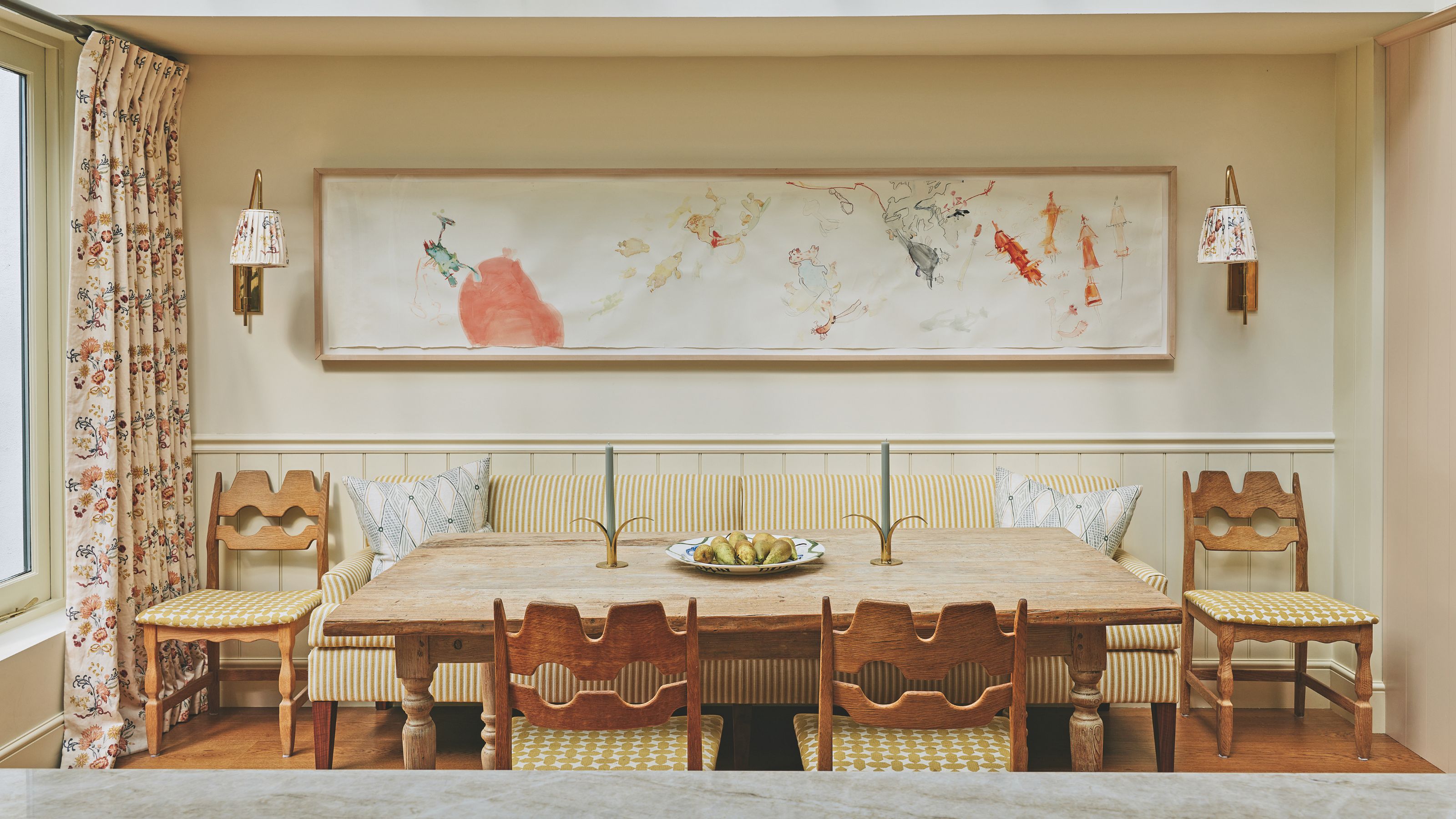 A dining nook with a long wooden table, a yellow striped banquette, and unique carved wooden chairs set beneath a large framed abstract sketch.