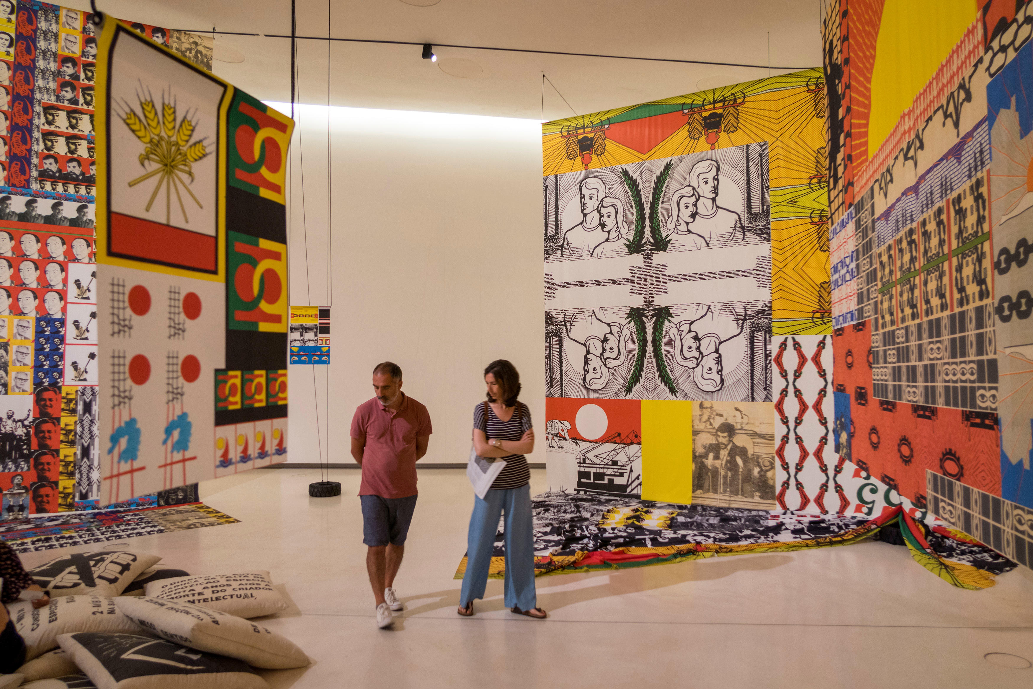 Two people walk through an art gallery. Large tapestries hang either side.