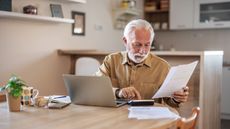 An older man uses a calculator and looks at paperwork at the kitchen table with a laptop in front of him.