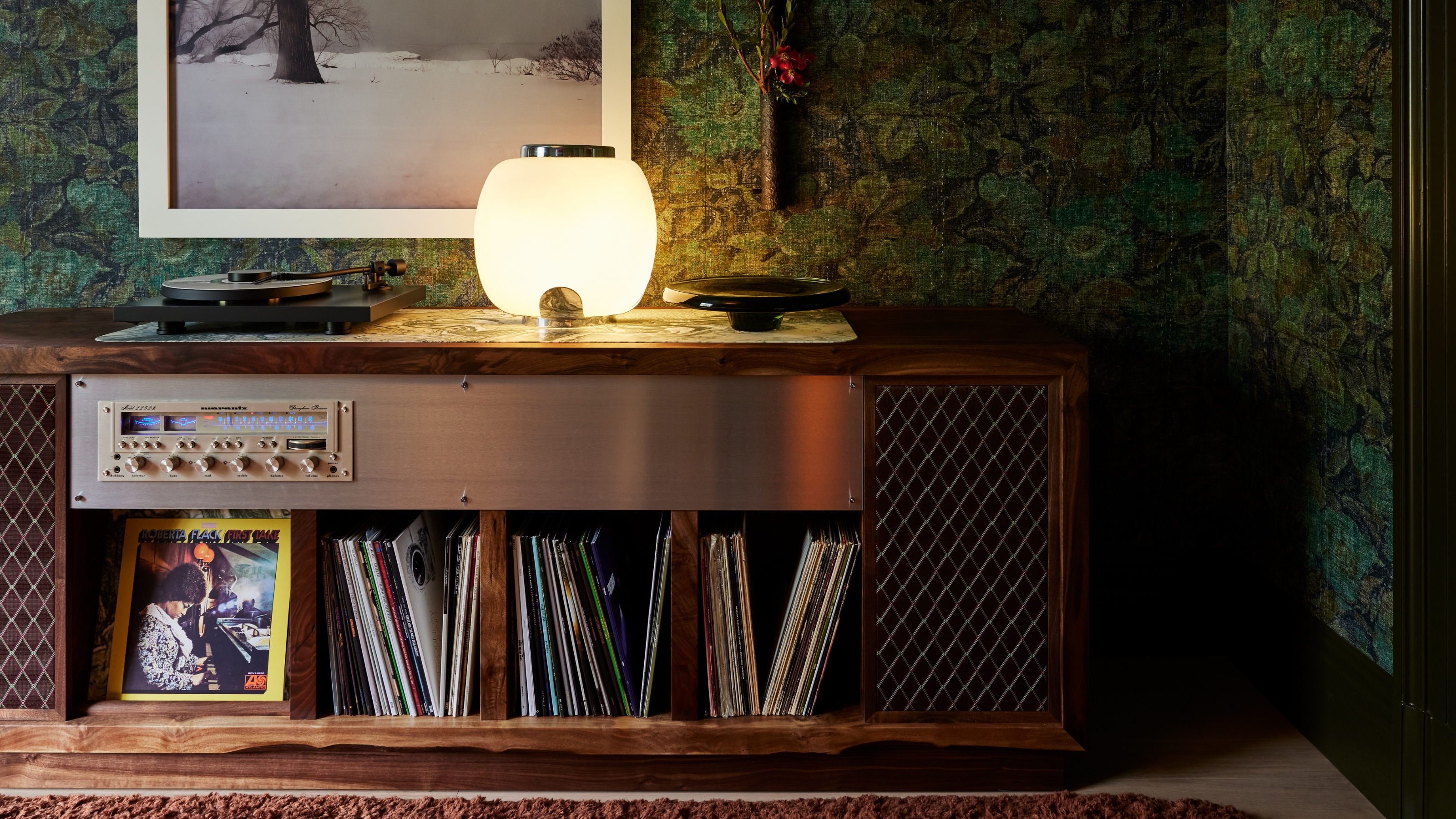 Image of a long, custom record player stand with built-in record storage. The walls of the room are a dark green wallpaper, and there is an orb light on the chest that&#039;s on.