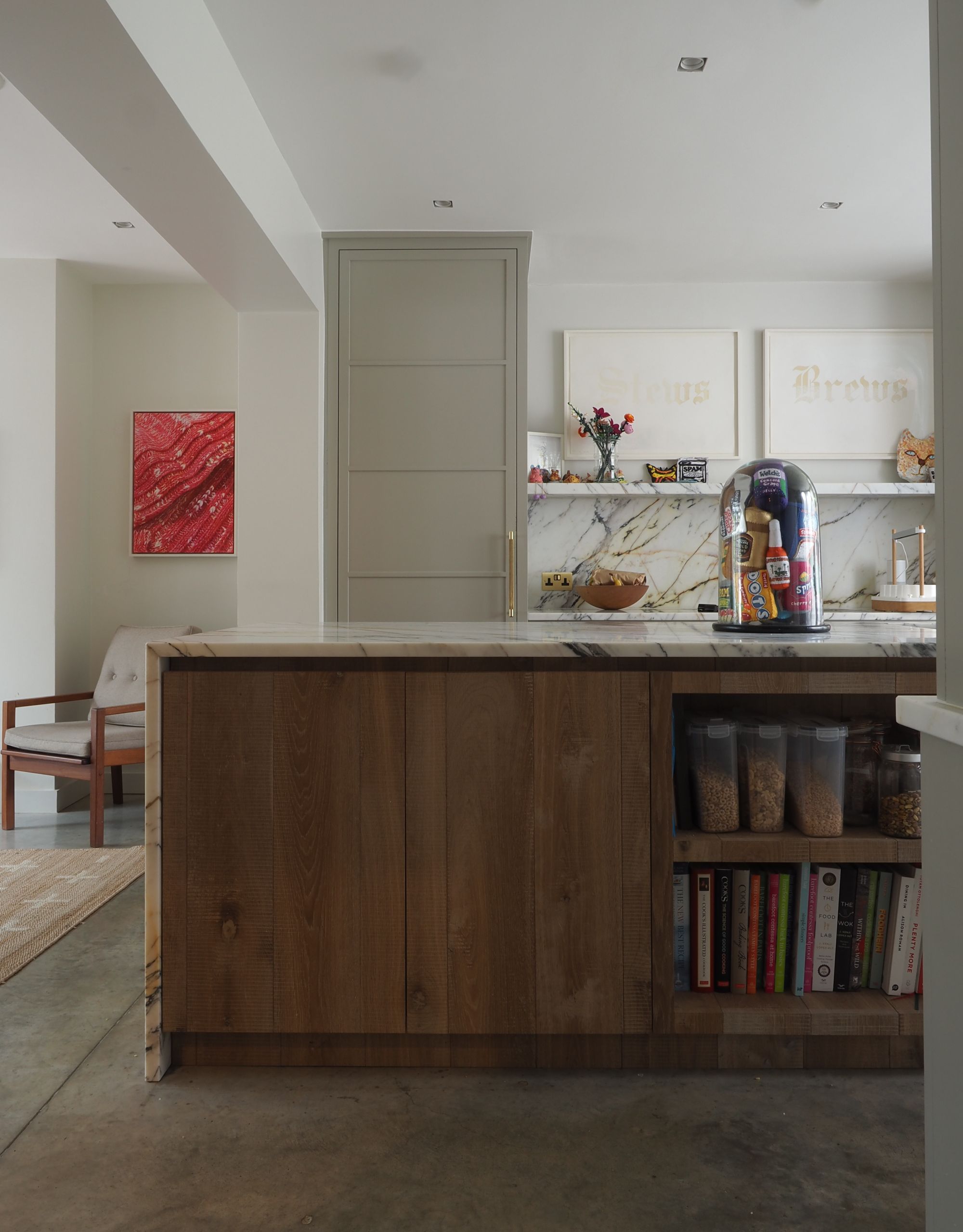 A modern kitchen with a large kitchen island with wooden cabinets and open shelves with a marble top and artwork on the wall and in a cloche on top of the island