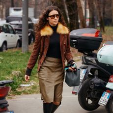 Model wears black turtleneck, leather jacket with fur trim, a pencil skirt, and tall boots. 
