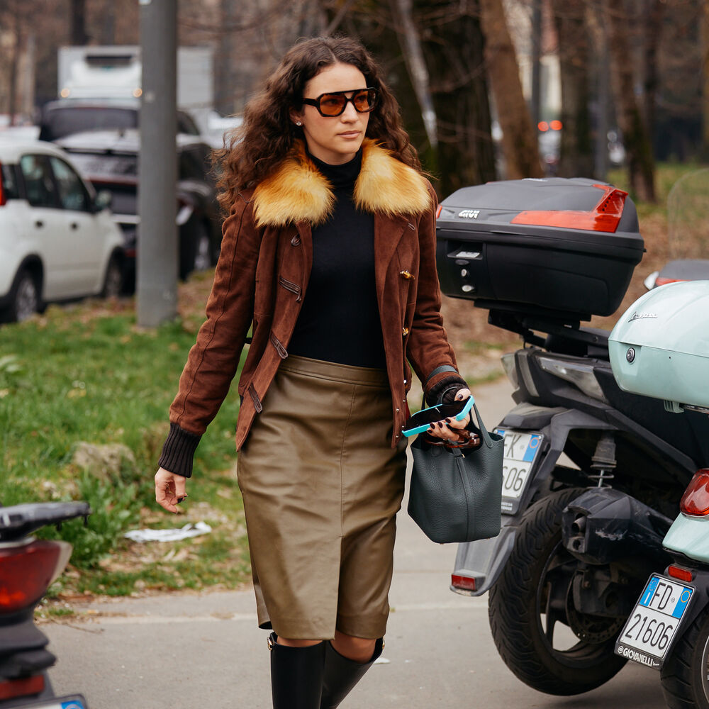 Model wears black turtleneck, leather jacket with fur trim, a pencil skirt, and tall boots. 
