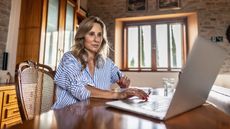 An older woman works on her laptop at her dining room table.