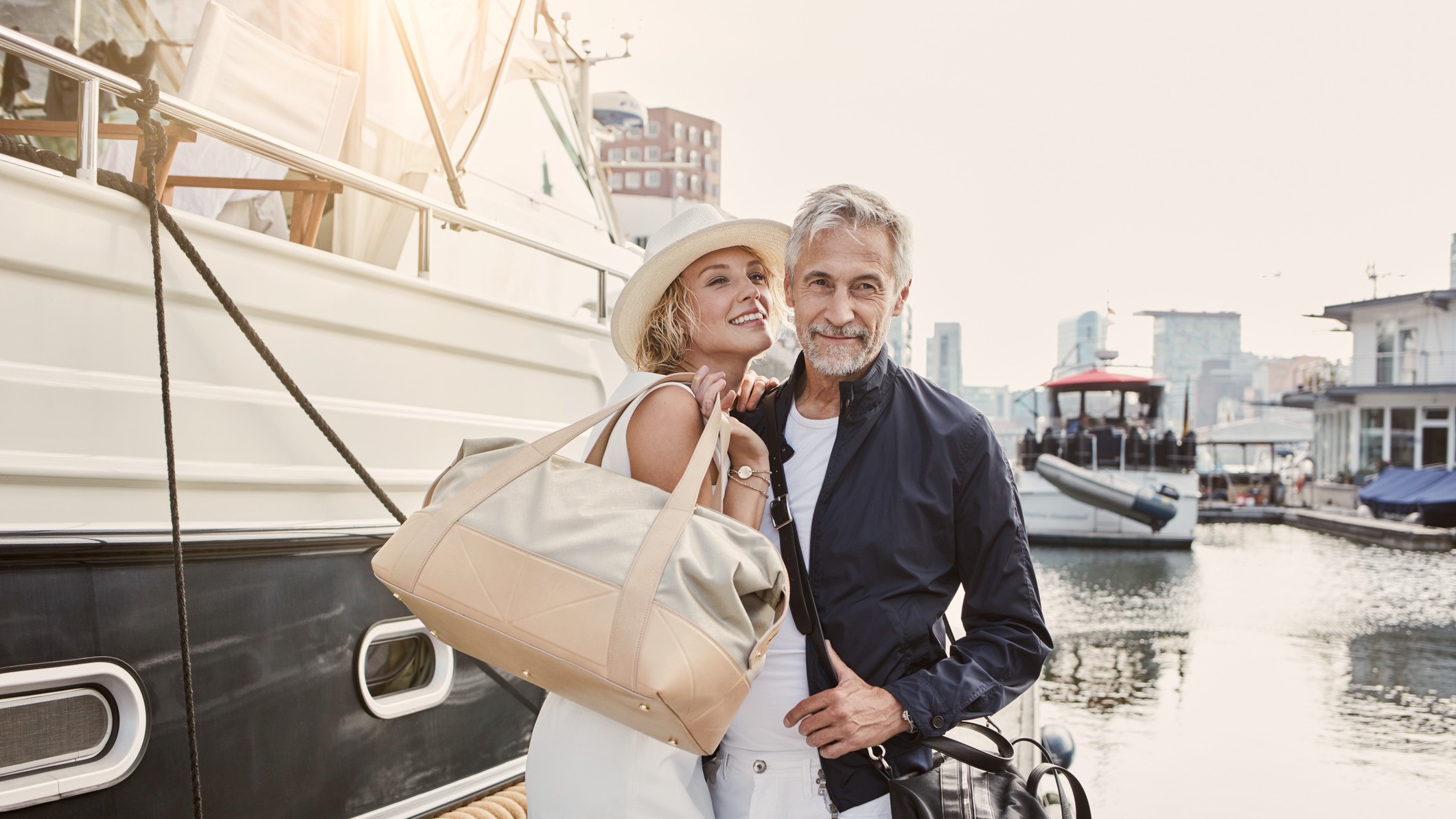 Older man and young woman standing with travelling bags on jetty next to yacht. They look insufferably smug.