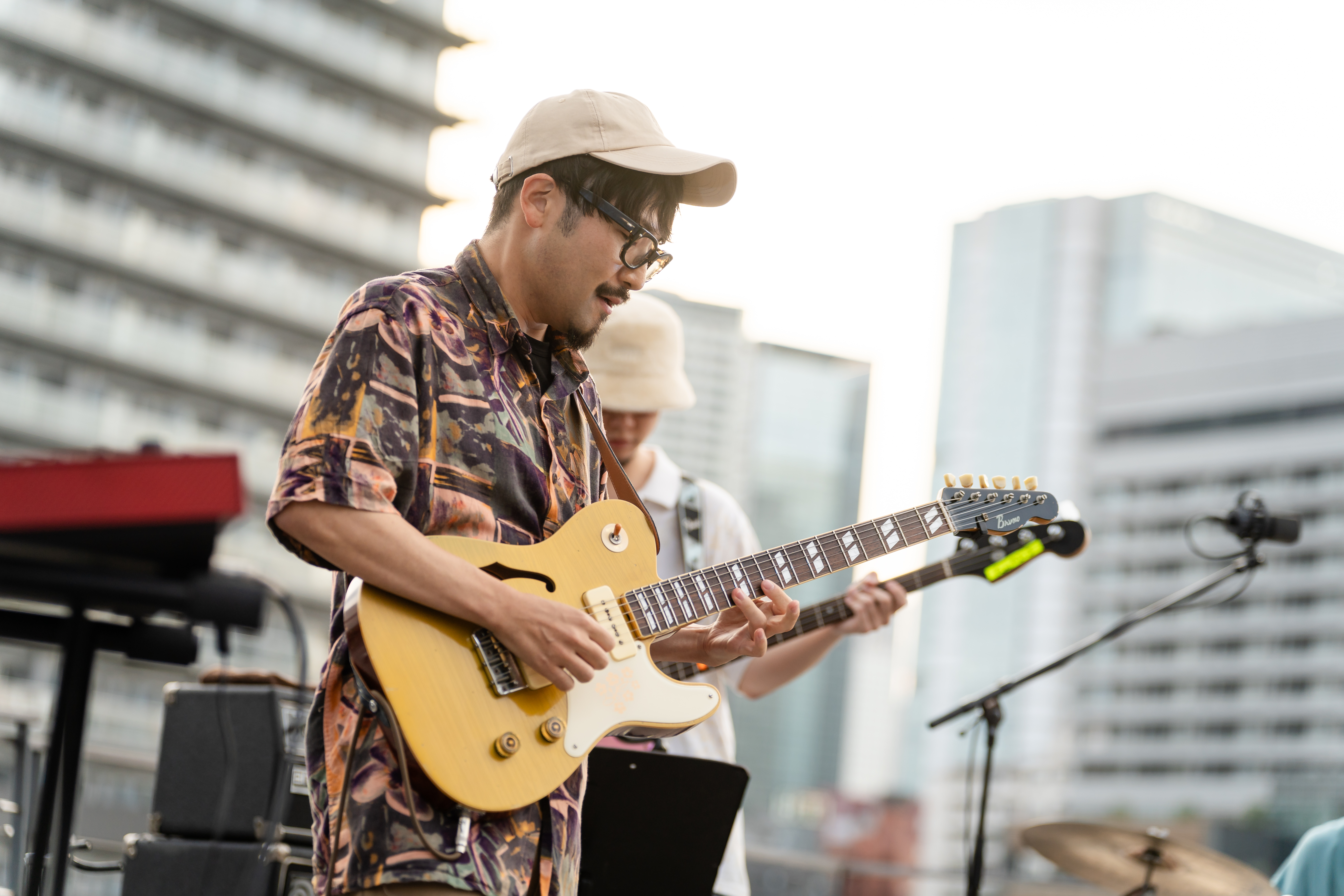 Toshiki Soejima playing guitar with a microphone, keyboards and blocks of flats in the background
