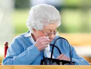 Queen Elizabeth wearing a blue sweater and wiping her eyes with a tissue with a black handbag in front of her