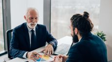 A boss smiles at his employee, a young man sitting across his desk.