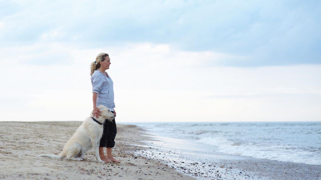 A woman looks off into the distance while standing on a beach with her dog.