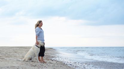 A woman looks off into the distance while standing on a beach with her dog.