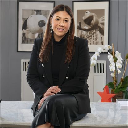 Nata Dvir, Chief Merchandising Officer at Macy’s, in a black dress and blazer sitting on a desk for Marie Claire's What I Wear to Work franchise.