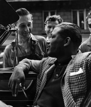 Black and white photo of US boxer, Sugar Ray Robinson sat in a car with three men surrounding his car, smiling.