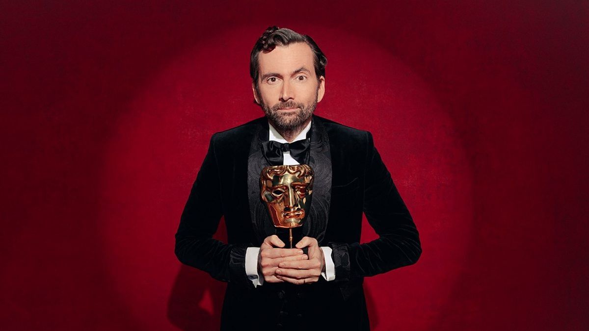 Host David Tennant in a black suit stands holding a golden BAFTA trophy, in a circle of light and standing in front of a red theatrical curtain.