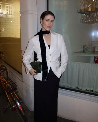 Norwegian Influencer Annabel Rosendahl poses outside a restaurant wearing a black skinny scarf, white blazer layered over a black dress, and a clutch bag