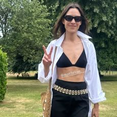 @alexachung walks along a grassy field wearing a black knee-length skirt with a black bra, white shirt, gold chain belt and brown rope sandals.