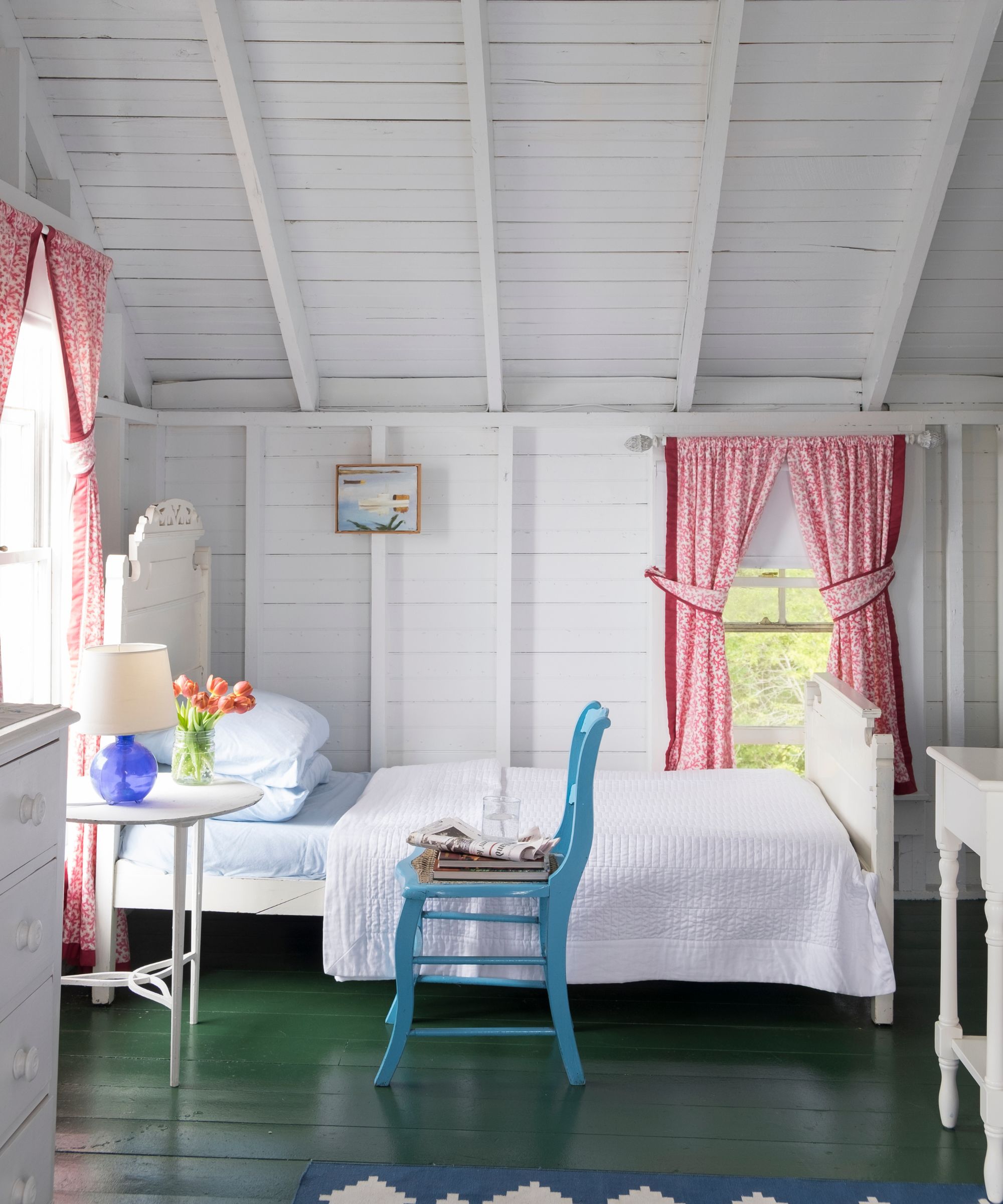 Bedroom with painted green wooden floorboards, white shiplap walls and red patterned curtains in Libby Cameron&amp;rsquo;s Maine coastal home.