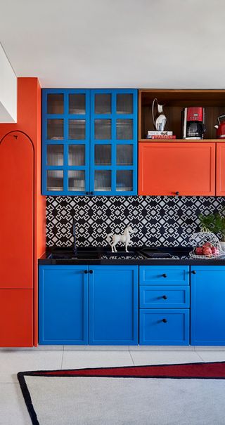 A small kitchen with red and blue cabinets and large prints on splashbackl