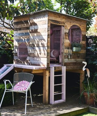 A DIY playhouse project on deck with pink painted ladder and slide with accompanying white chair and outdoor cushion
