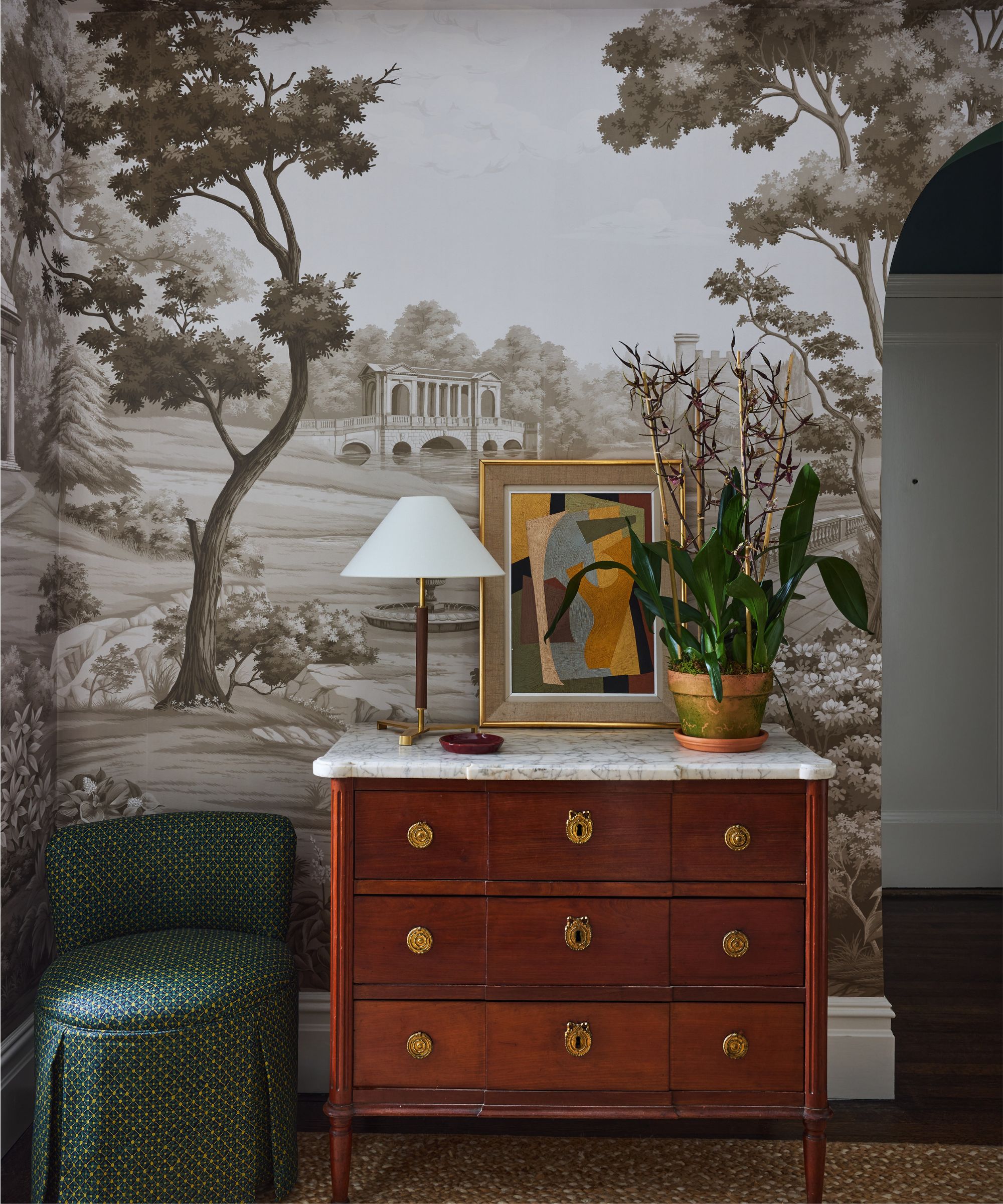 Entryway with mural landscape wallpaper in black and white, an antique mahogany dresser with a marble top, an antique painting, an Art Deco lamp sitting on top, and a skirted ottoman beside it.