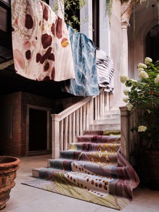 A series of pastel-shaded rugs decorated with abstract motifs hangs from the balconies and stone staircase of an old-style Italian palazzo.