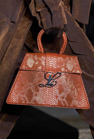 A snakeskin purse is shown in close-up photo of a model walking down the Luar spring 2025 runway show carrying a brown python handbag with a brown trench coat