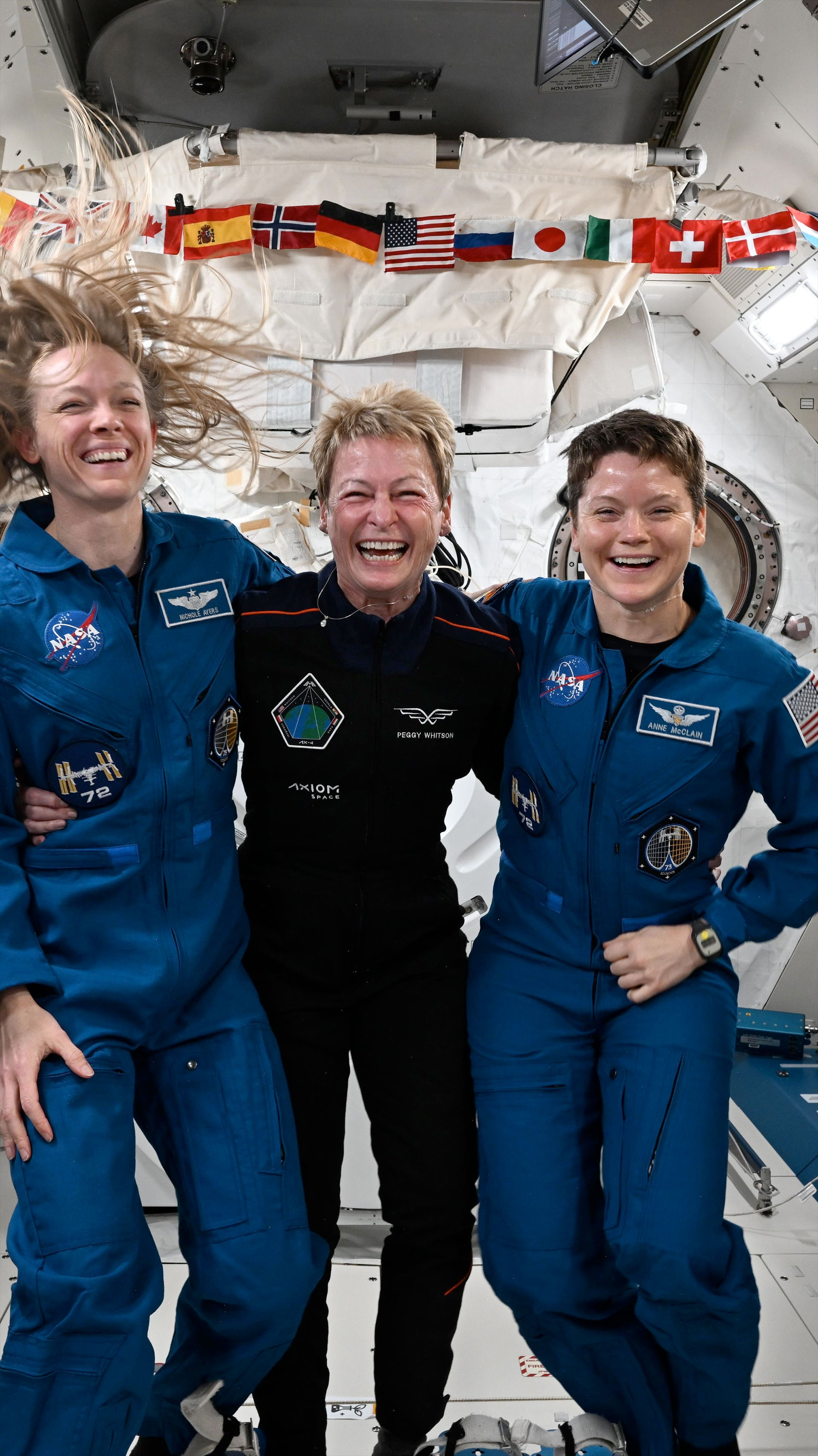 three women, two in blue flights and one in a black suit, pose and laugh together aboard a space station
