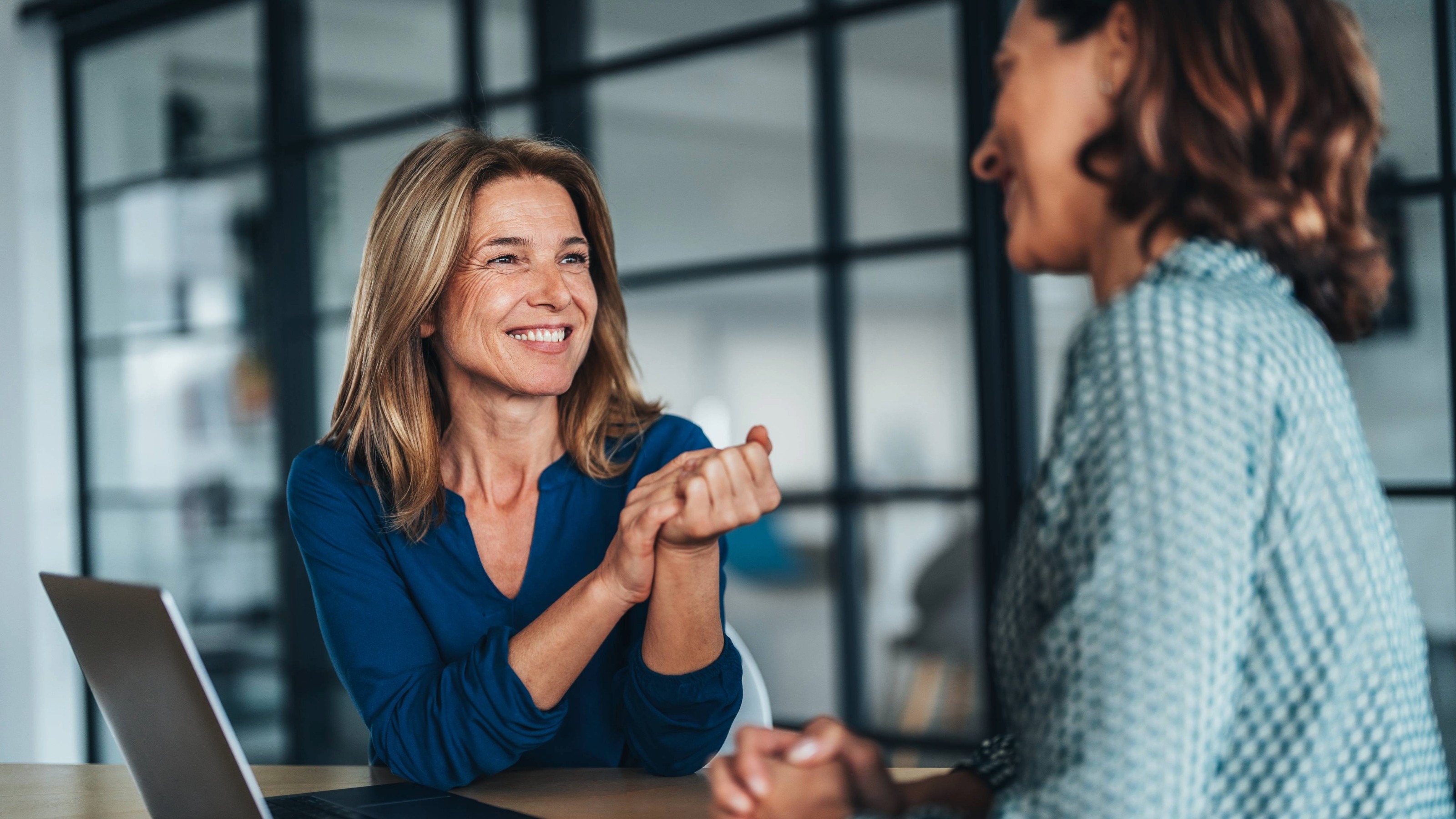 Two women having a friendly conversation at a desk. One woman talks and the other woman listens attentively while using a laptop.