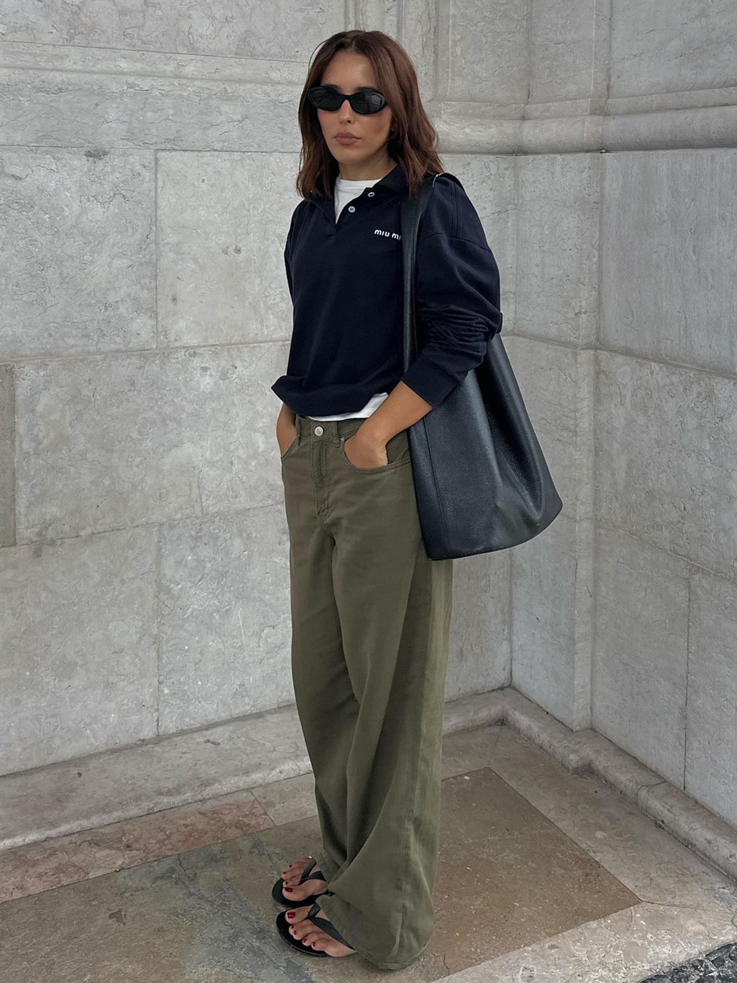style influencer Debora Rosa poses in a Miu Miu polo shirt layered over a white tee, a black leather tote bag, baggy green pants, and black flip-flop sandals