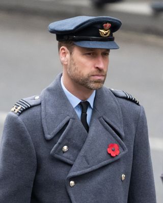 Prince William wearing a gray military coat and hat with a poppy brooch and a serious expression