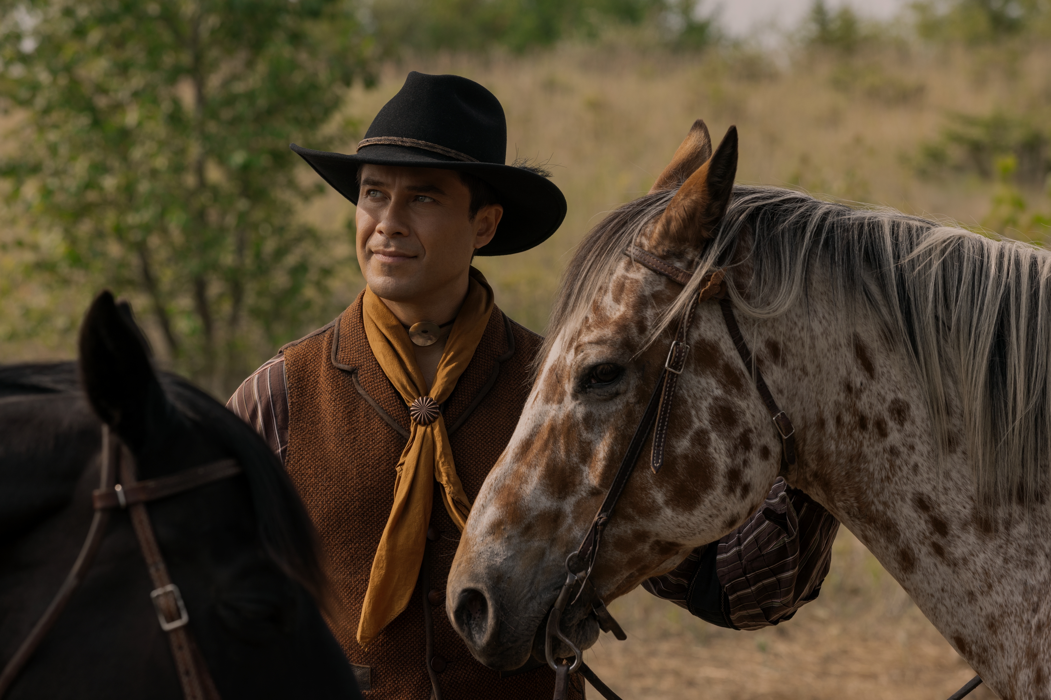 Actor Meegwun Fairbrother as Mitchell wears 19th-century clothing with a hat and a yellow scarf while standing outdoors between two horses, in Netflix's 'Little House on the Prairie.'