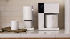 A white Fellow Aiden Precision coffee maker next to a white Fellow coffee grinder and a white mug on a white marble countertop.